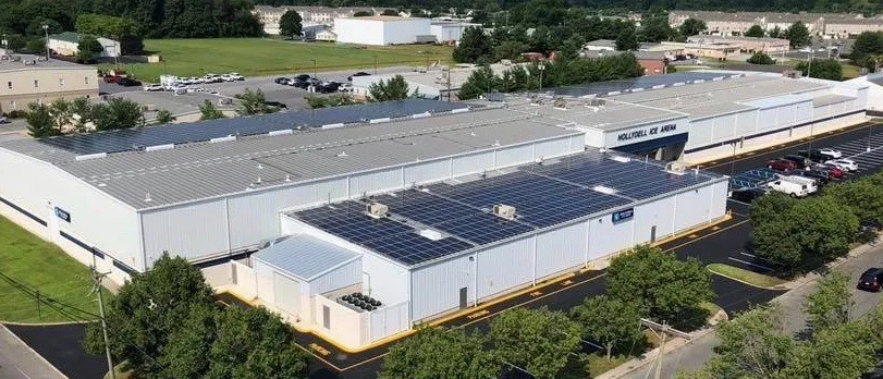 Aerial view of the low white metal-roofed complex with solar panels and "HOLLYDELL ICE ARENA" painted on the roof, parking lot and trees around it — Hollydell Ice Arena