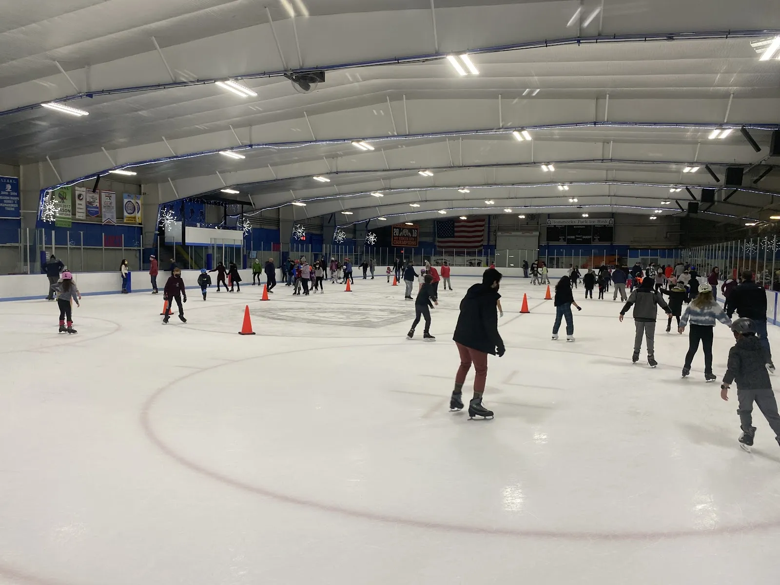 Una sesión de patinaje público en una pista amplia, con patinadores repartidos entre conos naranjas bajo un largo techo de luces y vallas azules y naranjas a lo largo del lado cercano. — Hommocks Park Ice Rink