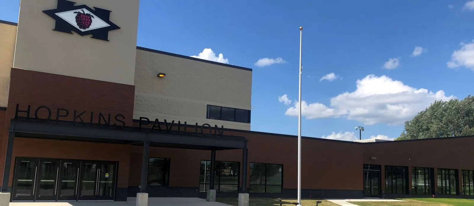 La entrada principal bajo las letras de HOPKINS PAVILION, con el logotipo de la frambuesa de Hopkins en la torre y un asta de bandera en el pasto, en un día soleado. — Hopkins Pavilion