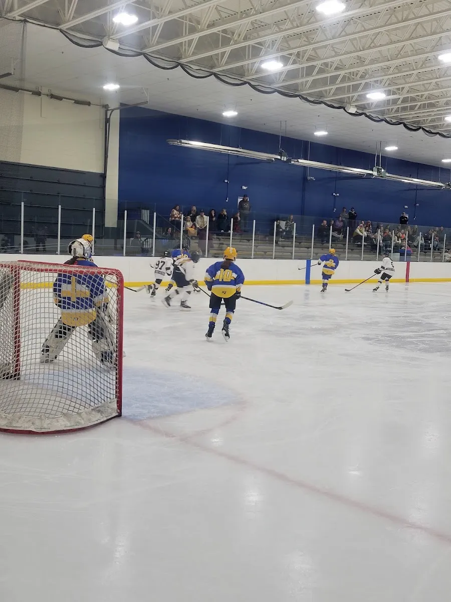 Un partido de hockey juvenil en curso en la pista de hielo: un portero frente a la portería a la izquierda, patinadores de azul y amarillo más allá, y espectadores en una grada baja bajo una pared azul y un techo de armadura de acero. — Hopkins Pavilion
