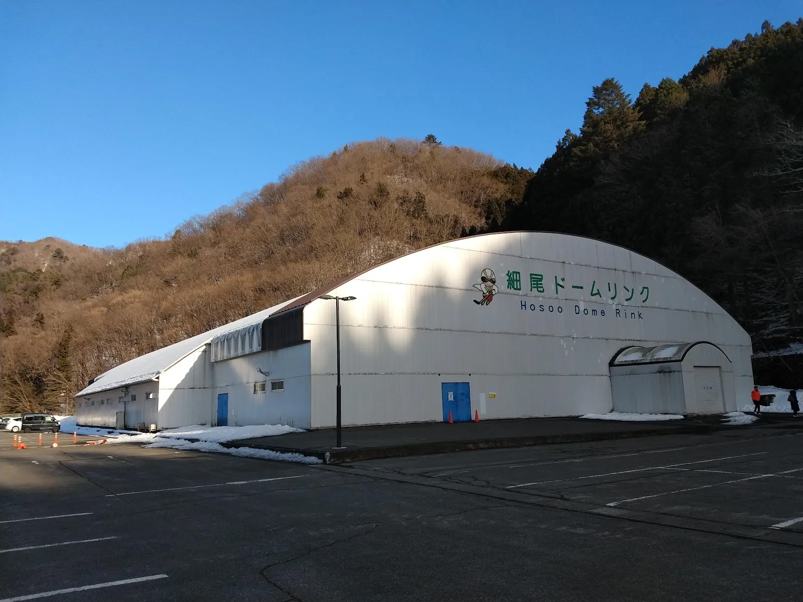 木々の茂る谷にある、Hosoo Dome Rinkの白いドーム。正面にリンクの名前とマスコットがあります — Furukawa Electric Hosoo Dome Rink (Nikko Hosoo Dome Rink)