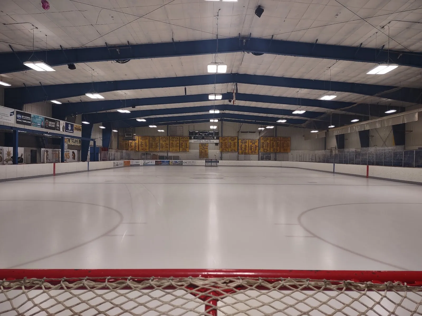 A wide empty sheet seen from behind a goal net, an arched blue-braced roof over the ice and banners along the far wall. — Howe Arena