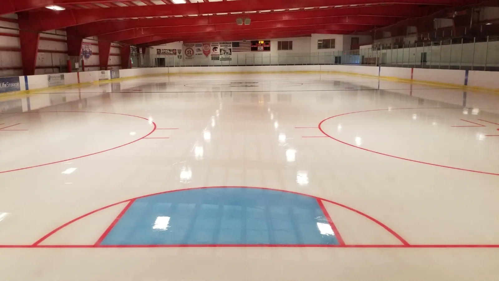 Fresh empty ice at Howell Ice Arena from the goal crease, the full sheet stretching away under red roof beams and banners — Howell Ice Arena