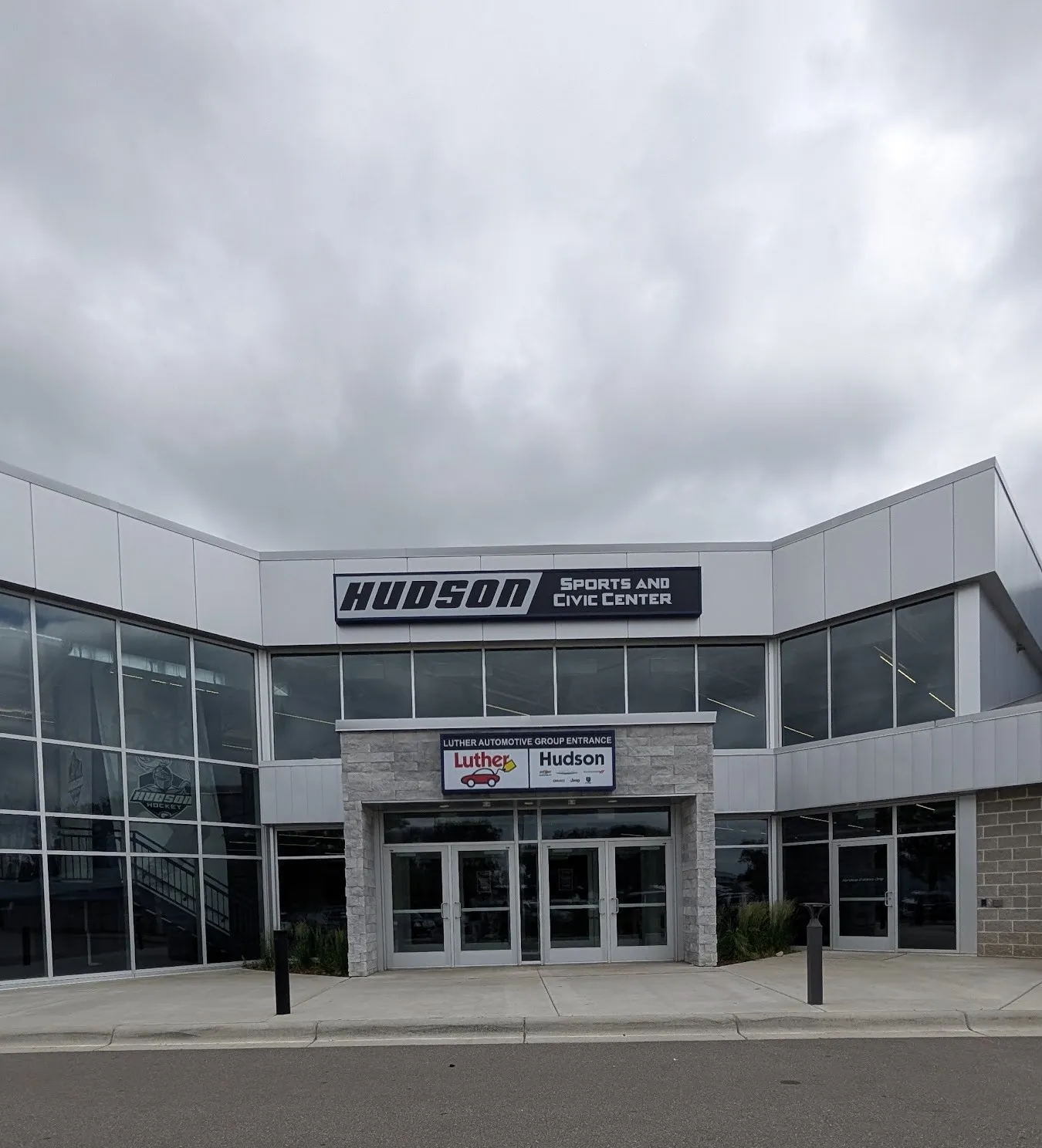 La entrada con frente de vidrio del Hudson Sports and Civic Center con el letrero de su nombre sobre las puertas, vista desde el estacionamiento bajo un cielo nublado — Hudson Sports & Civic Center