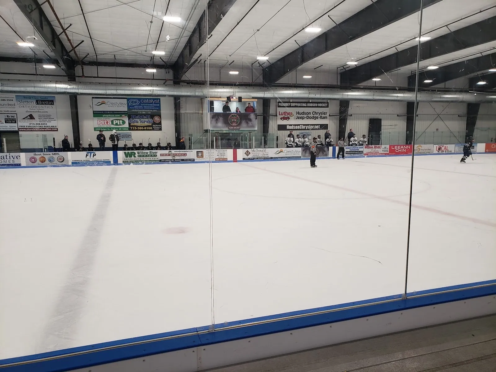 La pista vista a través de un vidrio transparente, más allá de un barandal azul, con unos cuantos jugadores a lo lejos sobre el hielo blanco bajo un techo de acero, con anuncios de patrocinadores en la pared del fondo. — Hudson Sports & Civic Center