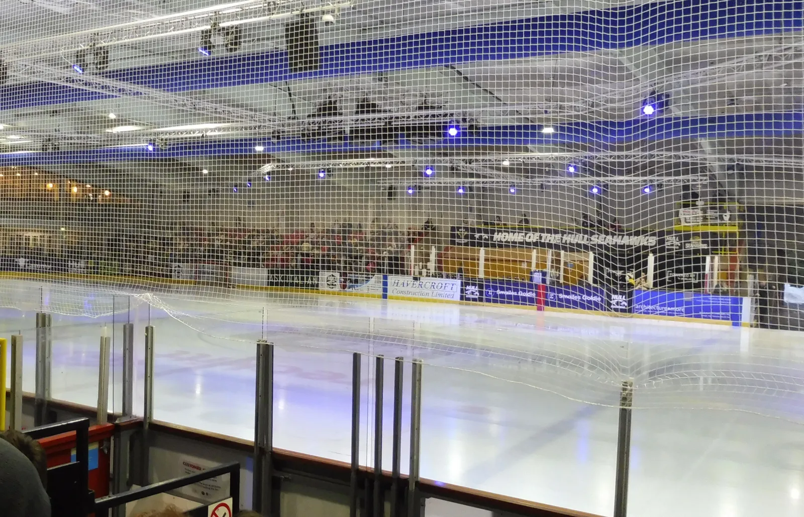 The Olympic-size ice seen through the safety netting before a hockey game, with a HOME OF THE HULL SEAHAWKS banner and sponsor boards along the far side (cropped). — Hull Ice Arena