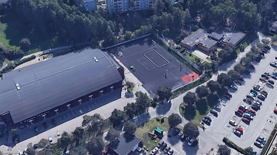 Aerial view of Husby ishall in Stockholm: the long dark-roofed hall beside its car park, a tennis court and the surrounding trees. — Husby ishall