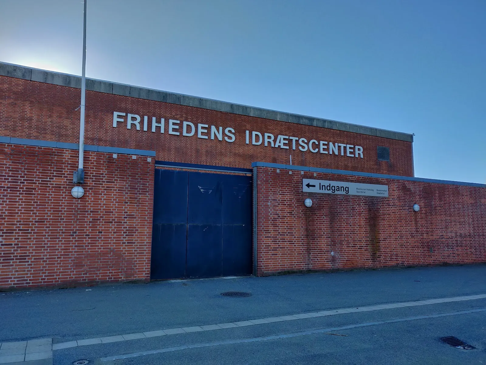 Den røde murstensmur på Frihedens Idrætscenter, idrætscentret med skøjtehallen, med navnet i stålbogstaver over en blå stålport og en pil mod indgangen. — Hvidovre Skøjtehal