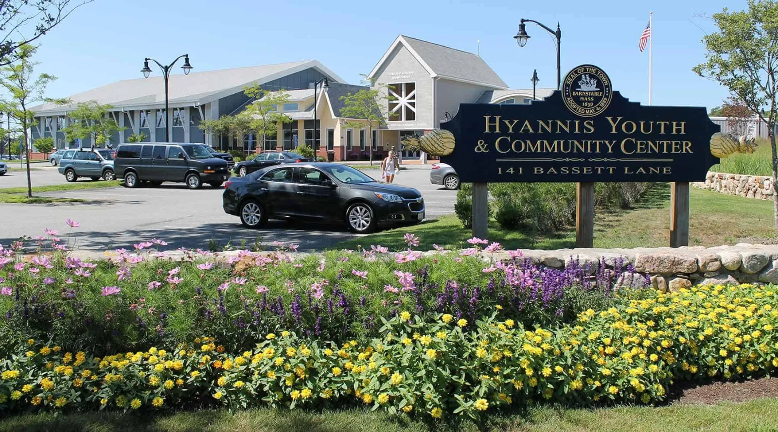 The entrance sign of the Hyannis Youth & Community Center, 141 Bassett Lane, with the building behind a flower bed. — Hyannis Youth & Community Center