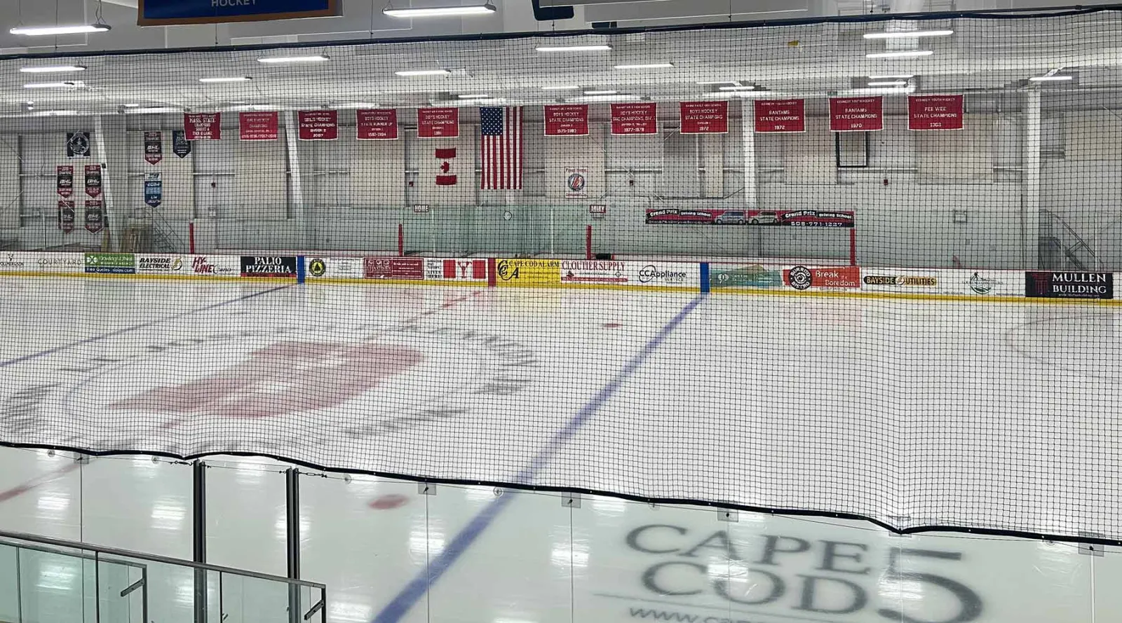 The ice of the Lt. Joseph P. Kennedy Jr. Memorial Skating Rink seen over the glass, with banners and sponsor boards on the far wall. — Hyannis Youth & Community Center