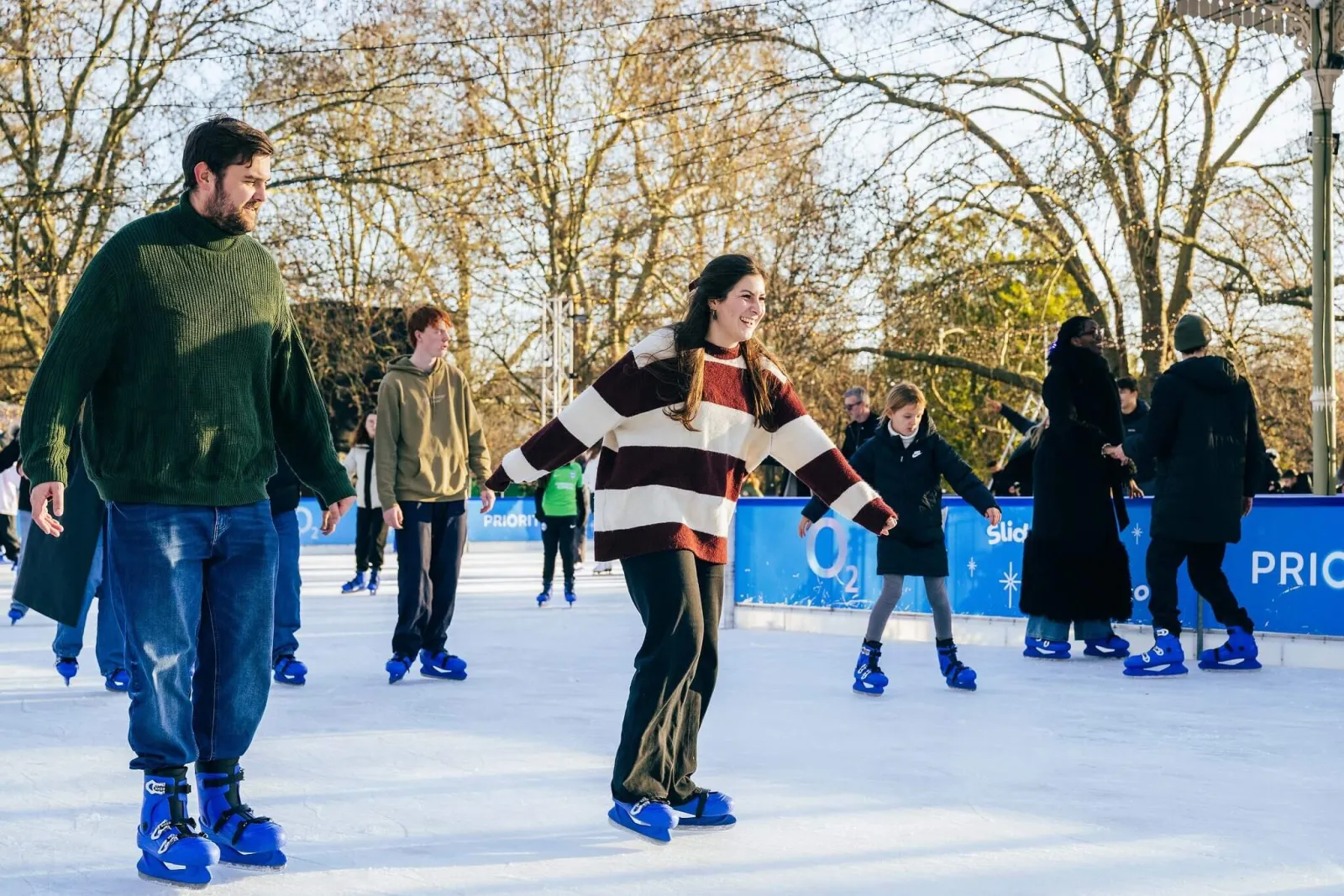 Skaters crossing the rink in daylight at Winter Wonderland, with bare trees behind and the boards in the background. — Hyde Park Winter Wonderland Ice Rink