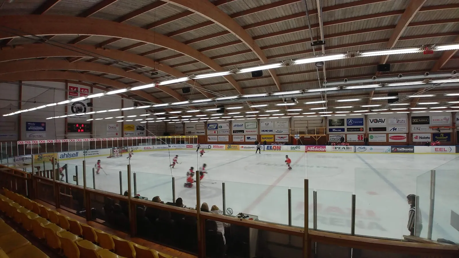 En bred vy över ishallen från sittplatserna längs långsidan, med ett välvt innertak i trä, några åkare och spelare på isen och sarg med reklampaneler. – ICA Maxi Arena Kumla