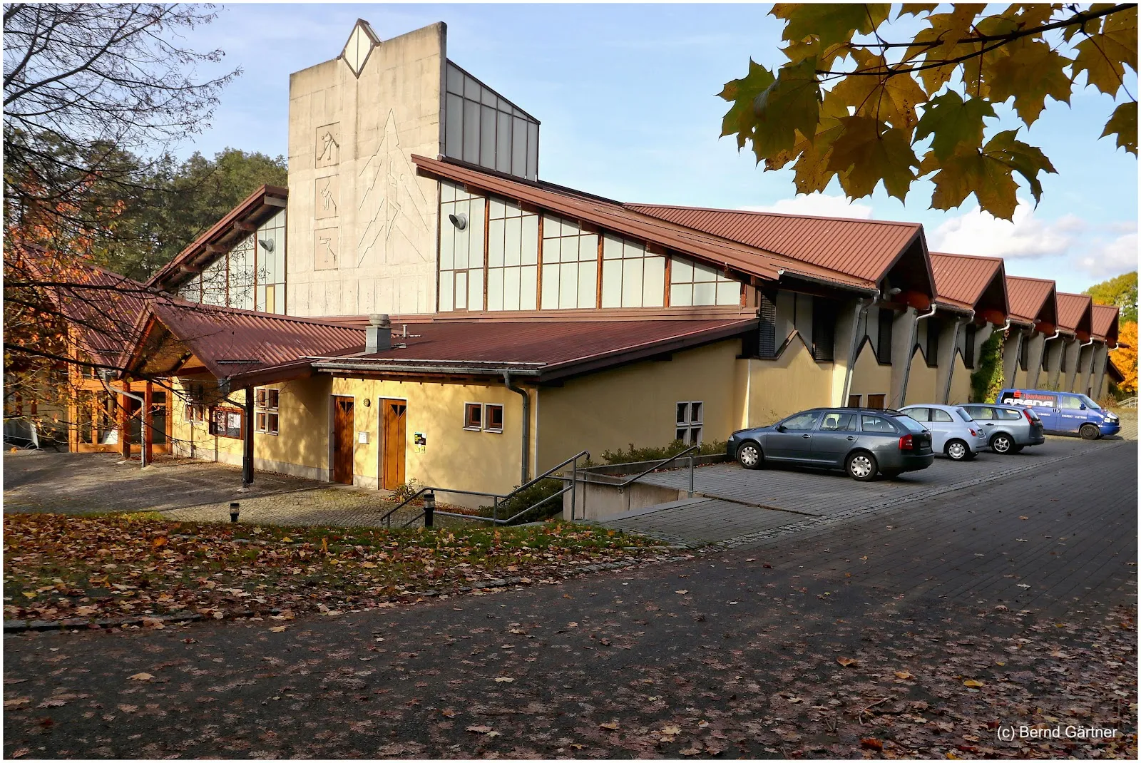 Das Gebäude der Eisarena von vorn: eine Halle mit Holzdach und heller Steinfront, einem steilen braunen Dach, dem Parkplatz und Herbstlaub. – ICE ARENA Jonsdorf