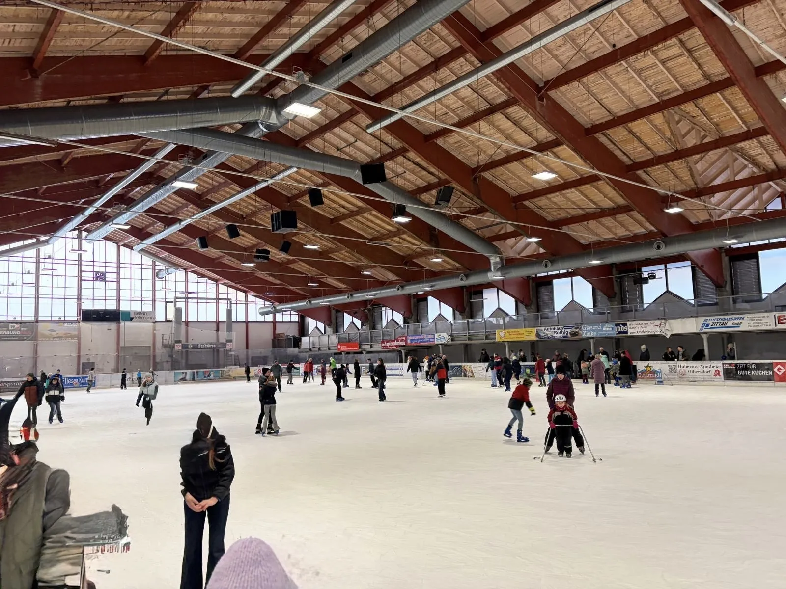 Die Weitansicht der Eishalle vom anderen Ende: die ganze Eisfläche mit kleinen Schlittschuhläufern in der Ferne unter dem Holzdach. – ICE ARENA Jonsdorf