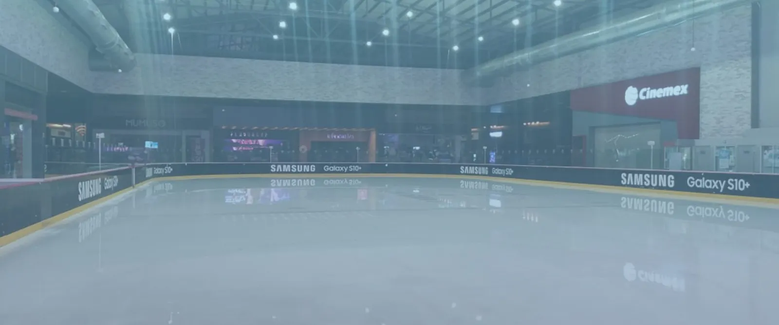 The empty ice sheet of the Puebla rink in a hall with sponsor boards along the wall. — Ice Arena Puebla