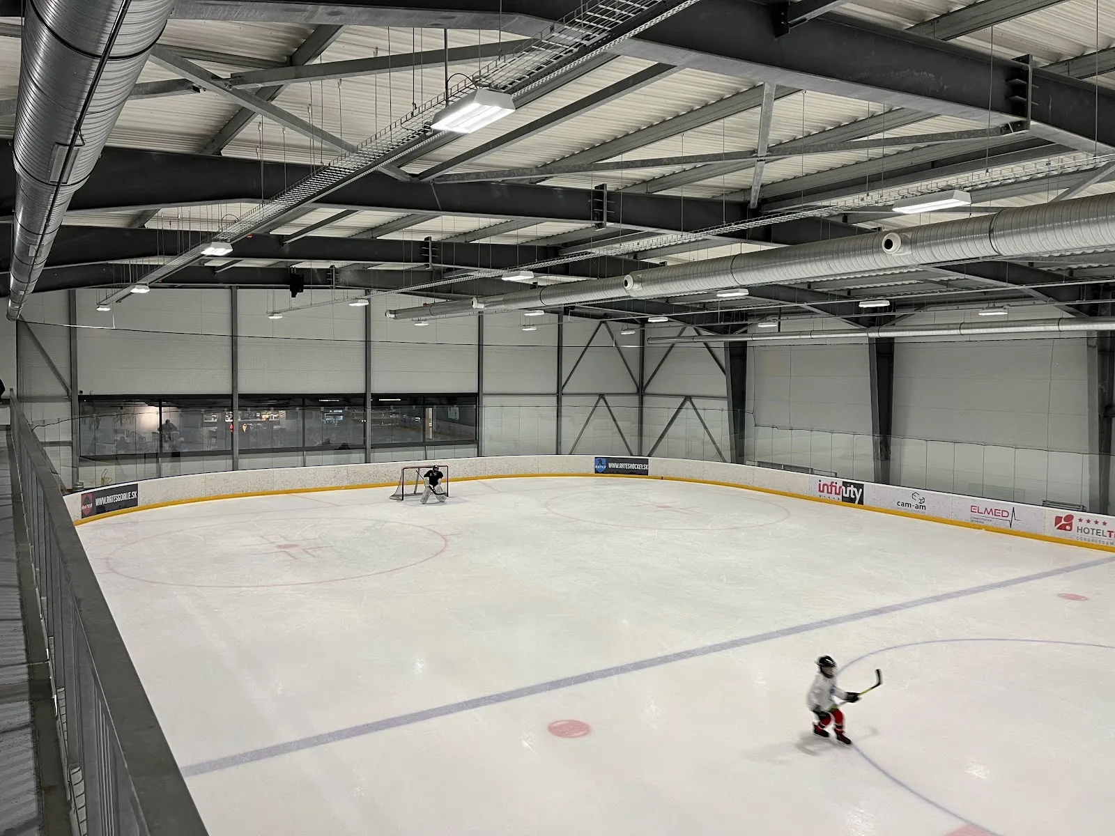 Raised view across the ICE ARENA Zvolen ice sheet with its boards, steel roof structure and a single distant skater. — ICE ARENA Zvolen
