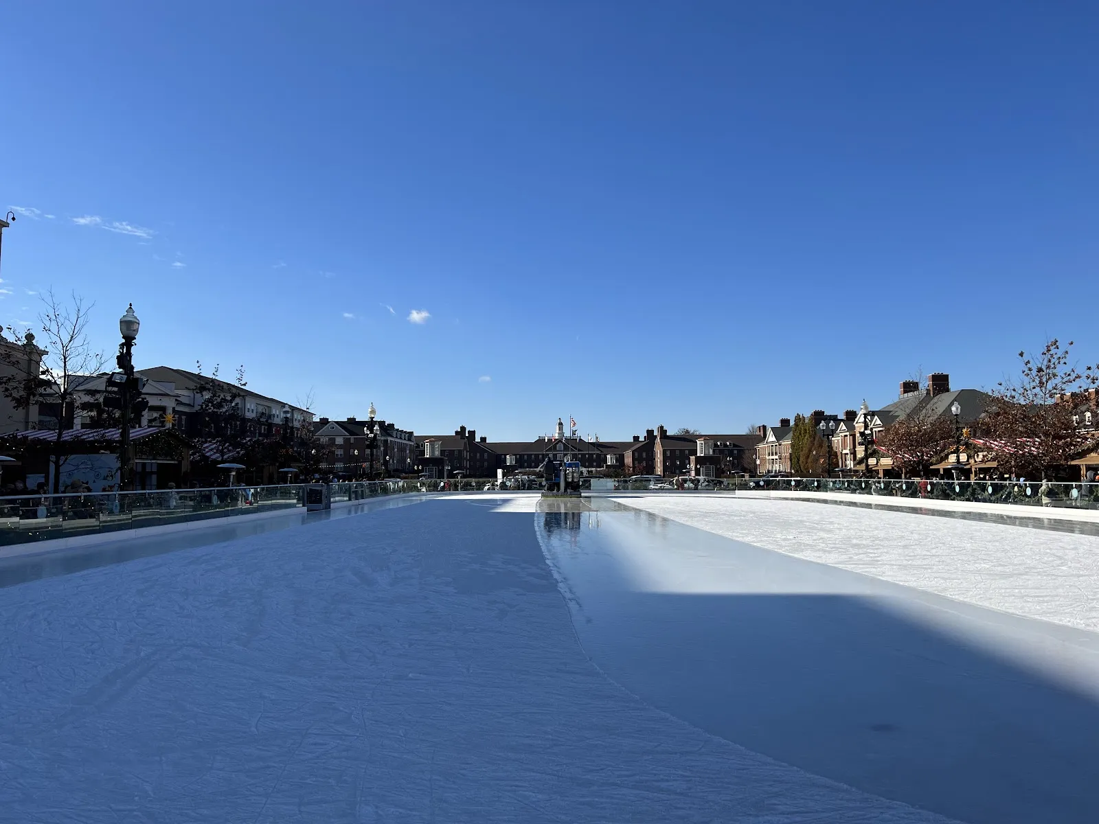The long outdoor rink at The Ice at Carter Green in Carmel under a clear blue sky, with the town square buildings beyond the rail — The Ice at Carter Green
