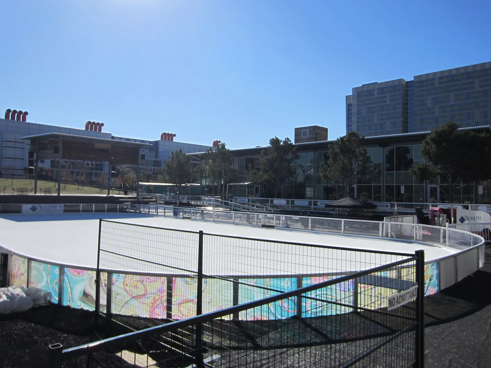 The skating rink at Discovery Green in downtown Houston in January 2012: a flat white ice surface with a fence and colourful boards, and park buildings behind — Ice at Discovery Green