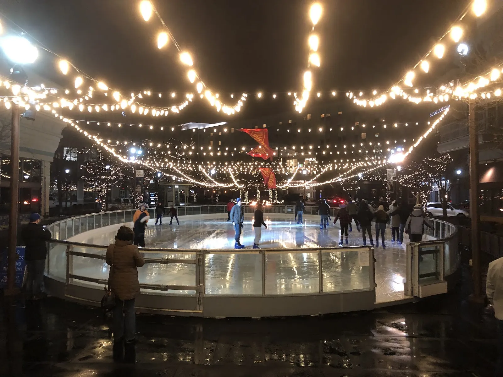 The oval rink at night inside its glass boards, under strings of lights, with skaters circling the ice. — The Ice at Park Place
