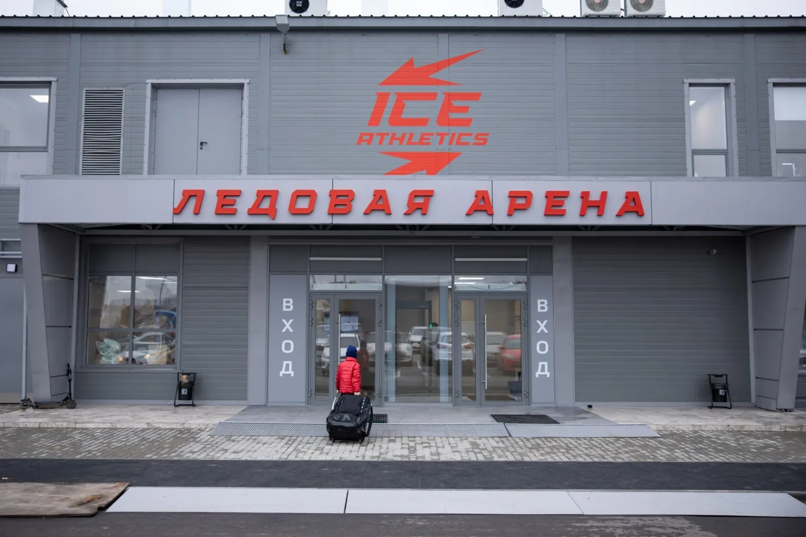 Серый фасад арены Ice Athletics с красным логотипом, надписью «Ледовая арена» над входными дверями и входящим с сумкой посетителем — Ice Athletics Arena Moscow