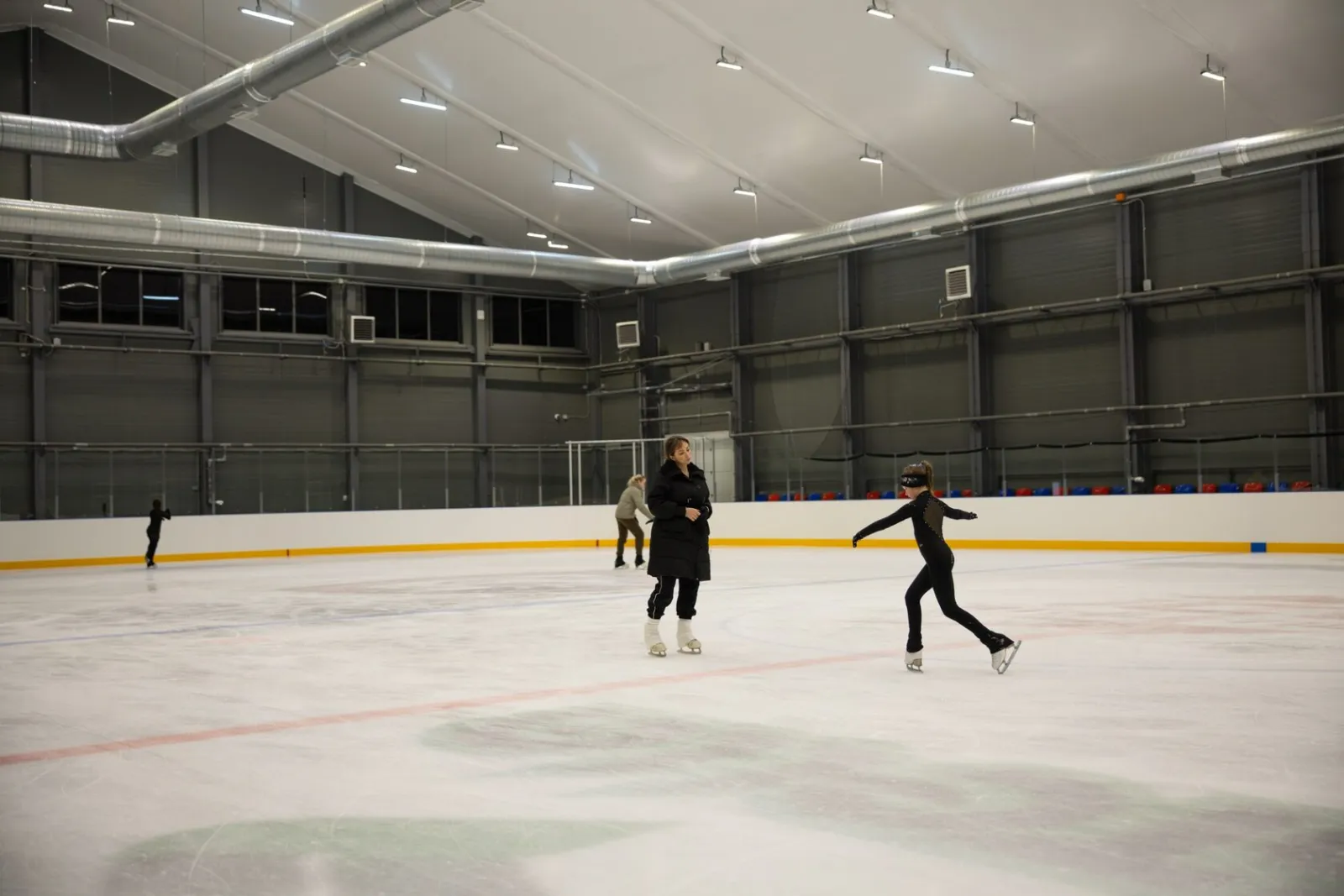 Несколько катающихся на широком льду поля Ice Athletics под освещённым потолком, вид от бортов — Ice Athletics Arena Moscow