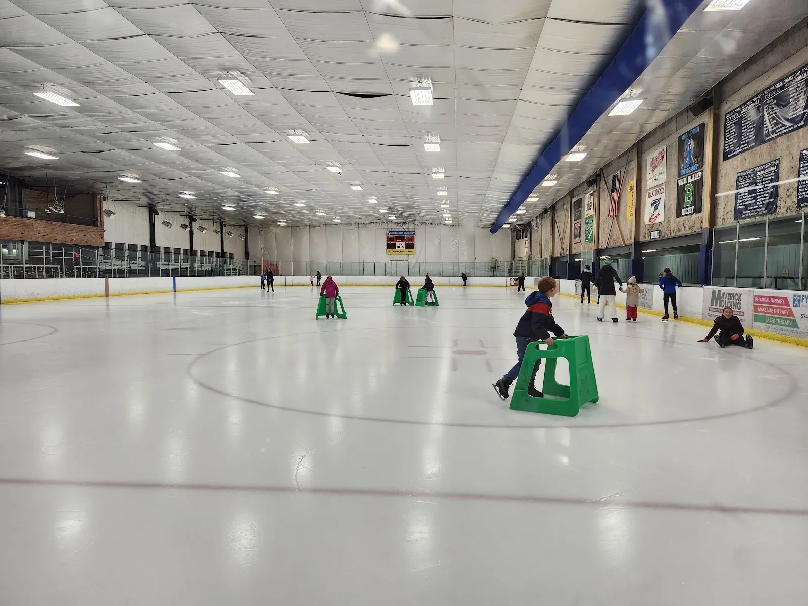 A wide empty-looking sheet with a few skaters and aid frames spread across the ice under a long ceiling of lights, blue and orange boards on the far side. — Ice Box Skating Rink
