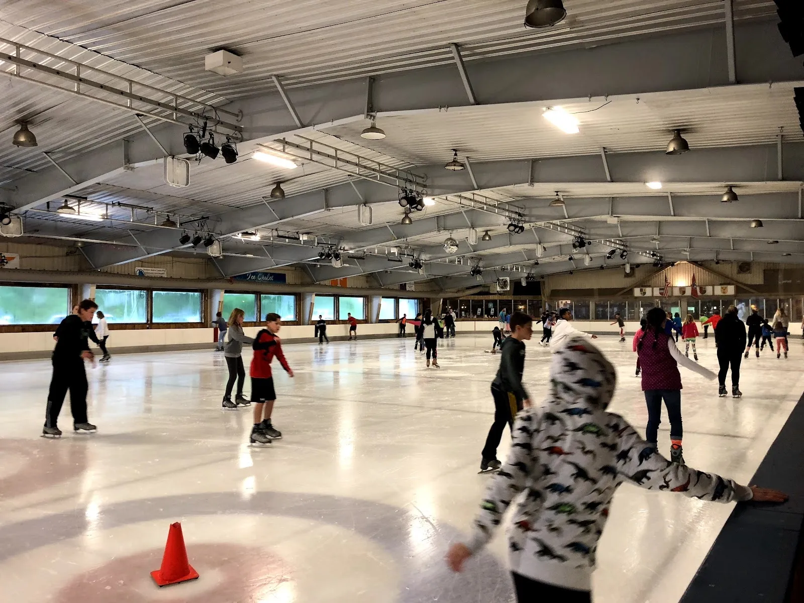 A public skating session on a wide sheet, skaters spread among orange cones beneath a pitched steel roof strung with international flags. — Ice Chalet