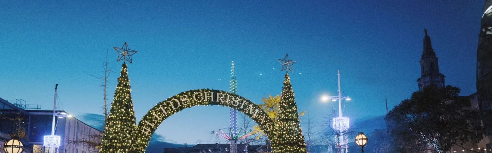 The glass-roofed Ice Cube marquee on Millennium Square in Leeds at dusk, with the lit Leeds Christmas arch and trees beyond. — Ice Cube at Christmas