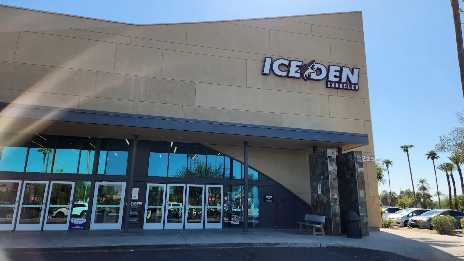 La entrada bajo el letrero de ICE DEN Chandler, una pared de puertas de cristal en la fachada color arena, con palmeras detrás del estacionamiento. — Ice Den Chandler