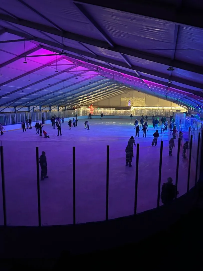 Das Eis unter dem weißen Zeltdach des ICE DOME in Mainz, violett und rosa beleuchtet, mit klein über die Eisfläche verteilten Schlittschuhläuferinnen und Schlittschuhläufern – ICE DOME Messepark Mainz