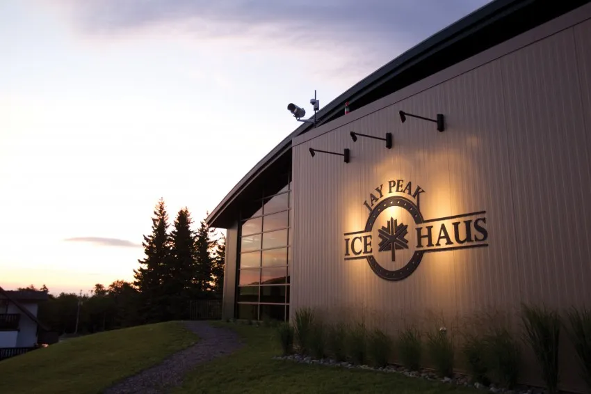 El muro metálico de la arena al anochecer con el logotipo iluminado de Jay Peak Ice Haus, un muro de vidrio en el extremo y abetos junto al camino. — Ice Haus Arena