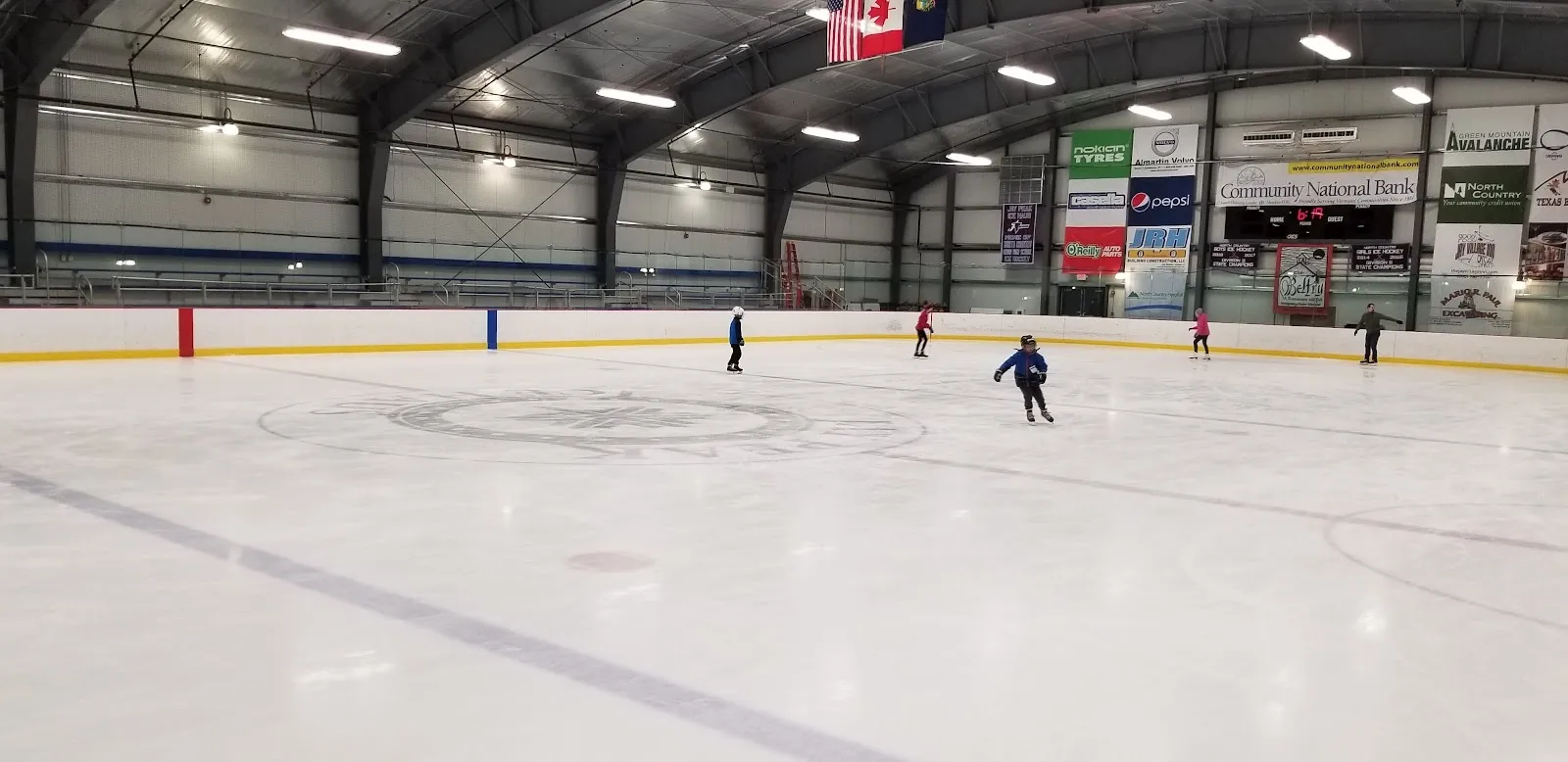 Una vista amplia a través del hielo con un patinador a lo lejos, en un largo recinto con estructura de acero y anuncios de patrocinadores en la pared del fondo. — Ice Haus Arena