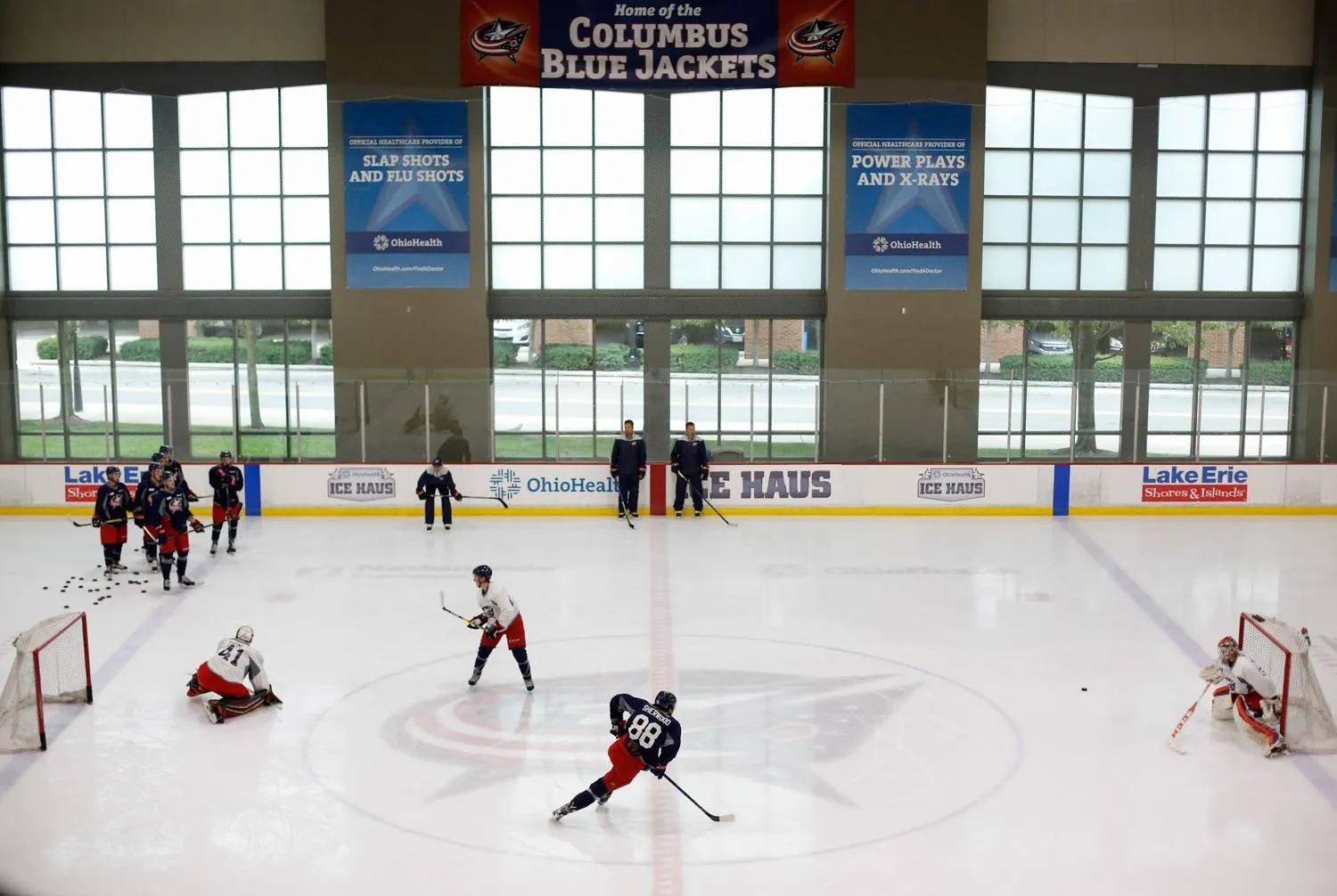 A faceoff in front of the Blue Jackets-banner wall, players spread across the ice below the arena's tall window grid. — OhioHealth Ice Haus