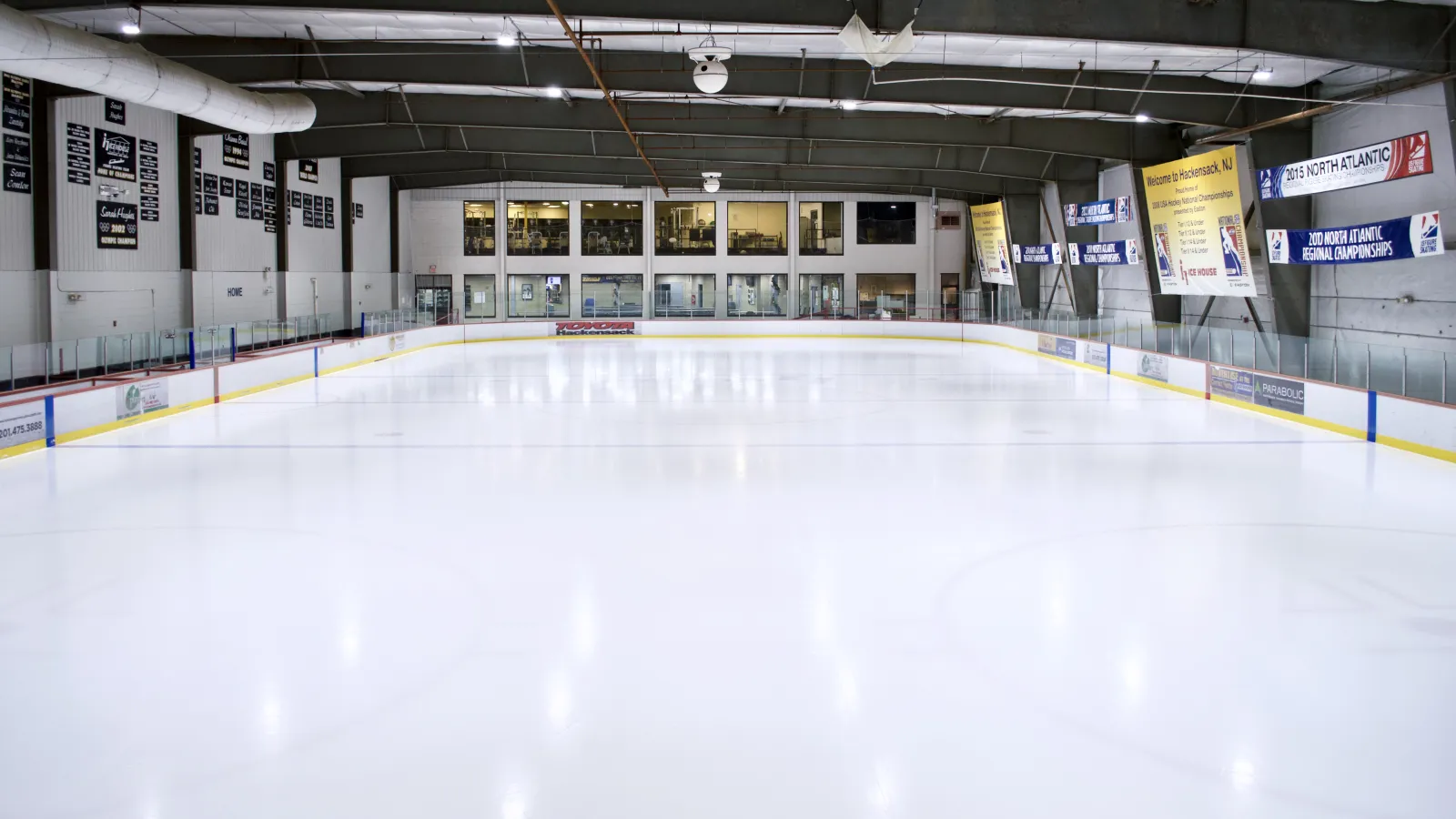 An empty, freshly resurfaced NHL-size sheet with blue-trimmed dashers, sponsor banners and the upstairs viewing windows at the far end. — Ice House of New Jersey