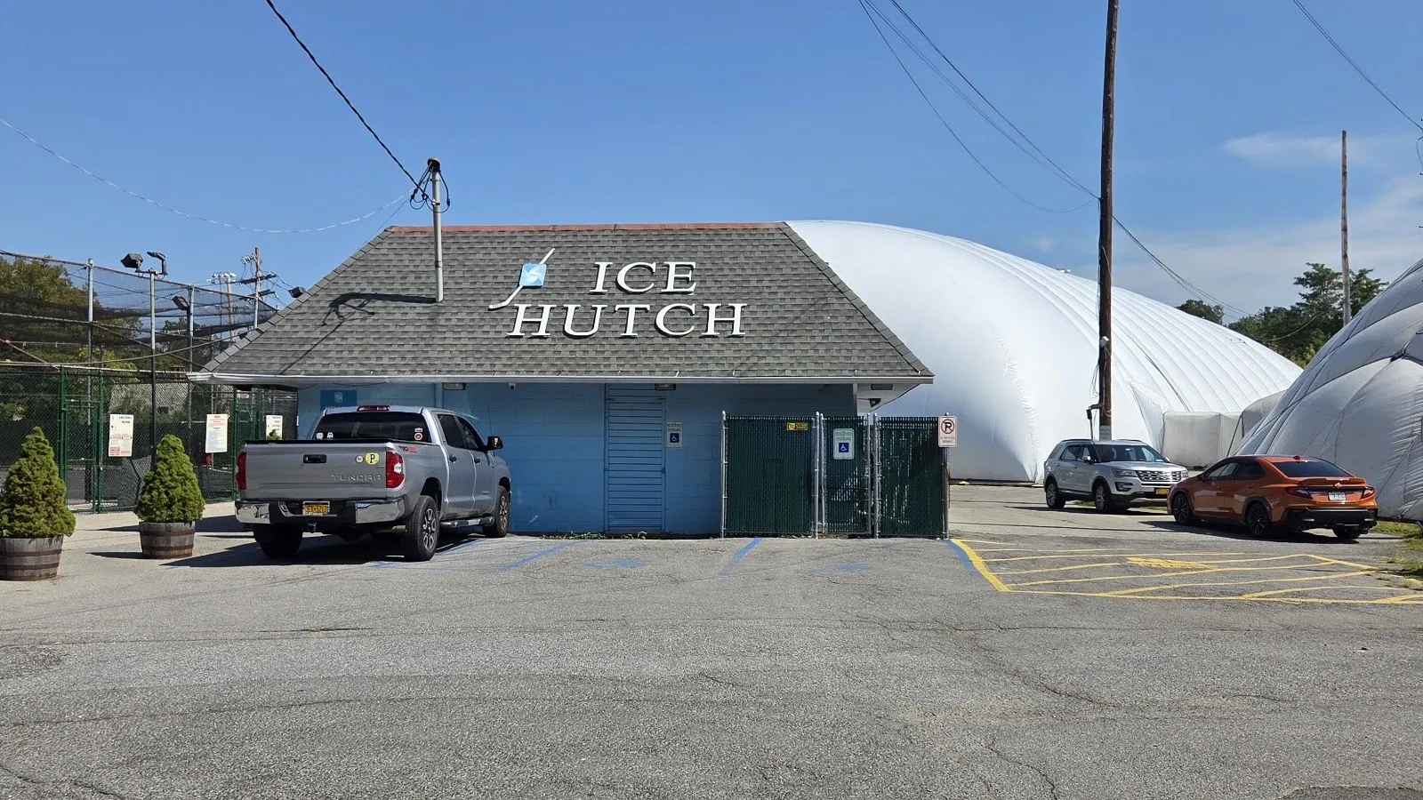 El edificio de entrada de Ice Hutch, con las letras ICE HUTCH en su techo gris y el domo inflable blanco de la pista que se eleva detrás. — The Ice Hutch