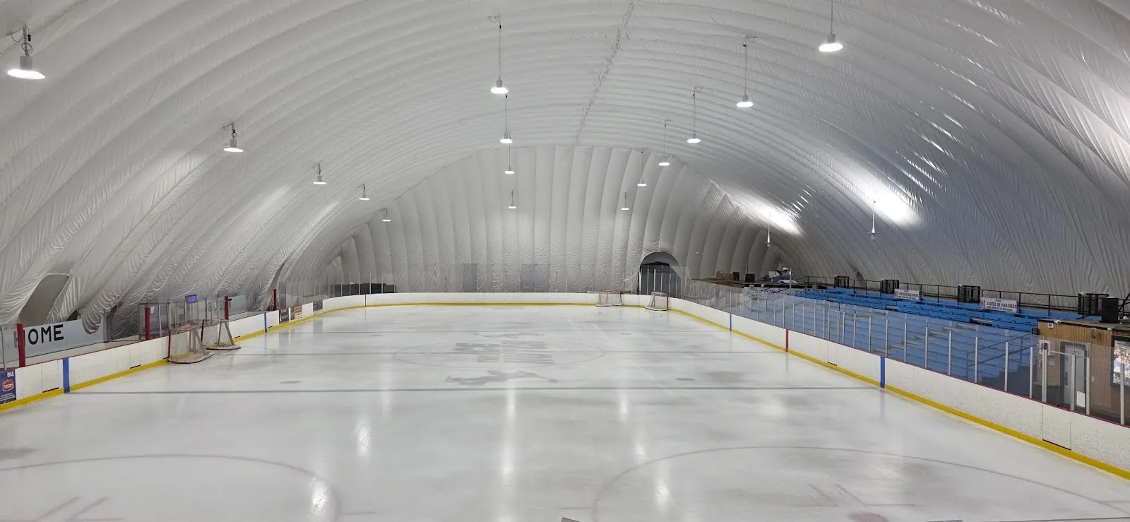 La pista vacía bajo el domo blanco inflable, con el borde azul de las bardas, una portería en el extremo del fondo y luces colgando del techo curvo. — The Ice Hutch