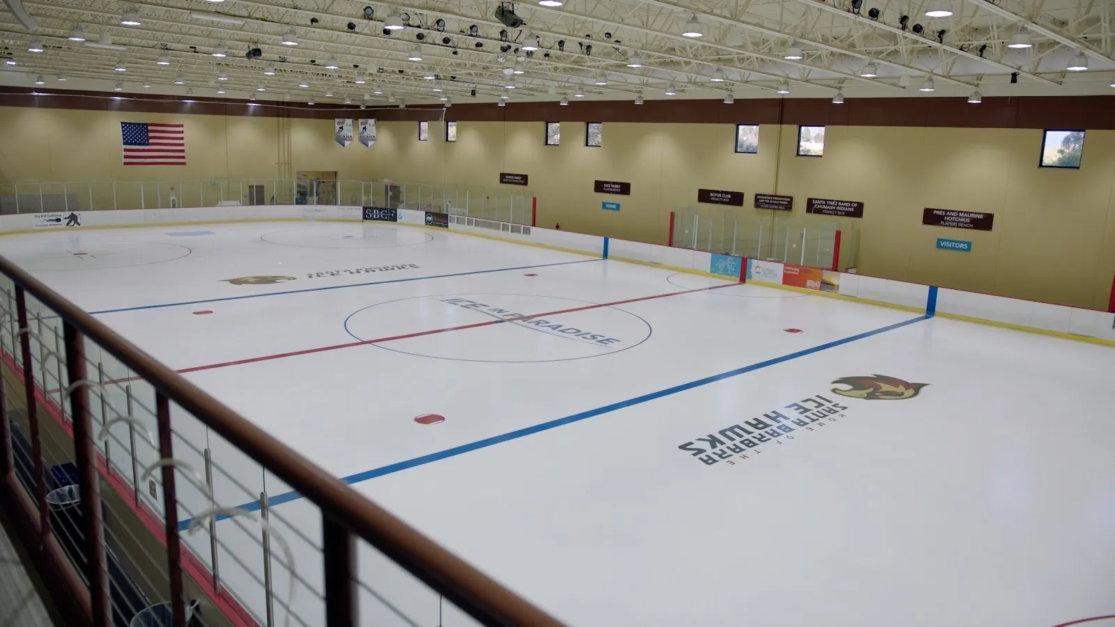 Una pista vacía vista por encima de un barandal oscuro, con líneas rojas y azules sobre hielo blanco bajo un techo de luces y el nombre Ice in Paradise en las vallas. — Ice in Paradise