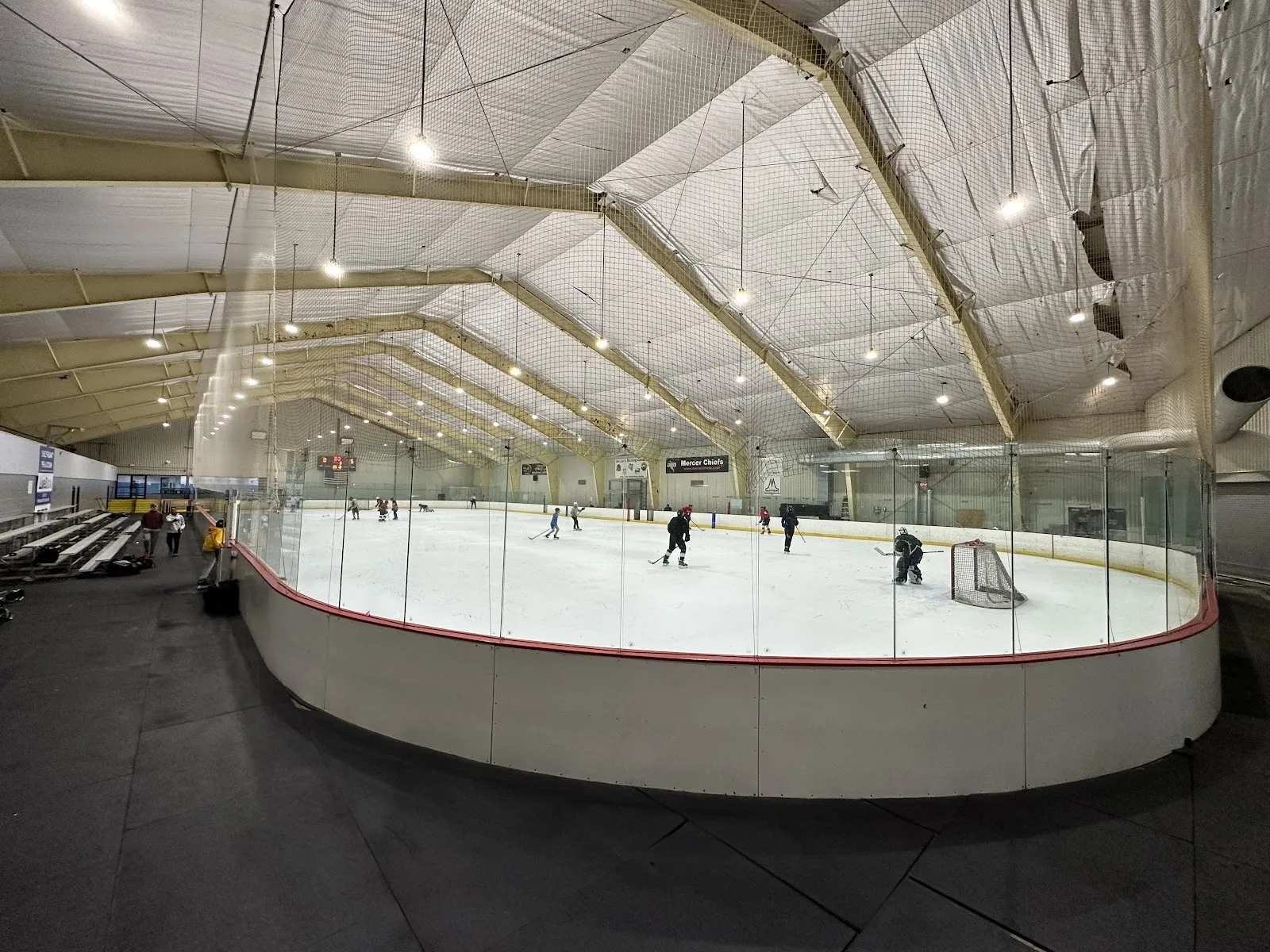 A wide view over the curved boards, a few skaters spread across the sheet under a white gabled roof with steel braces and hockey goals at the ends. — Ice Land Skating Center