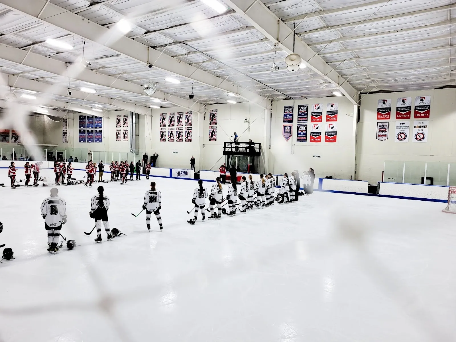 Two lines of players facing each other across the sheet before a game, a steel roof and rows of banners along the far wall. — Ice Line