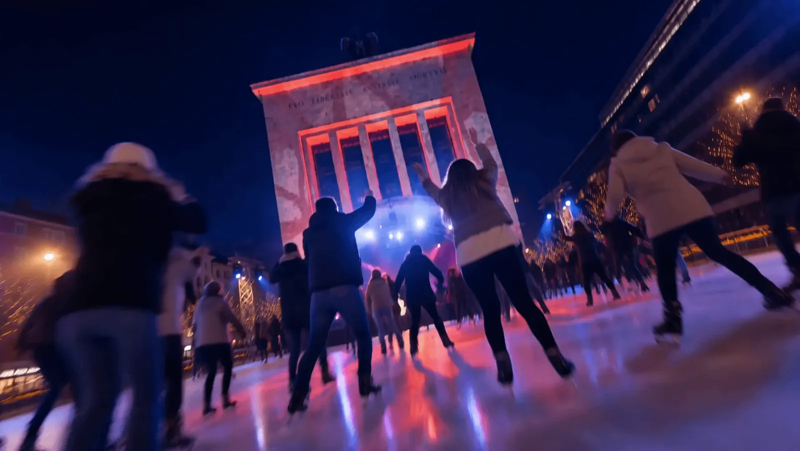 Skaters seen from behind on the Ice Magic rink at Landhausplatz in Innsbruck at dusk, with the lit memorial arch in the background. — ICE MAGIC Innsbruck (Landhausplatz & Bozner Platz)