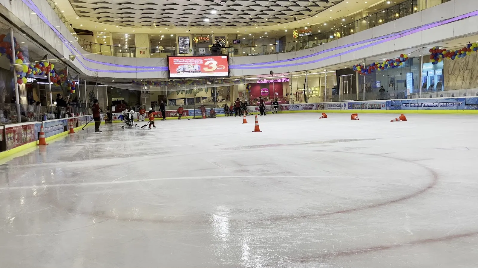 The ice sheet of ICE NAKA ARENA Rangsit inside the Future Park mall, with a few skaters and training cones on the ice and the mall atrium around the rink. — ICE NAKA ARENA Rangsit