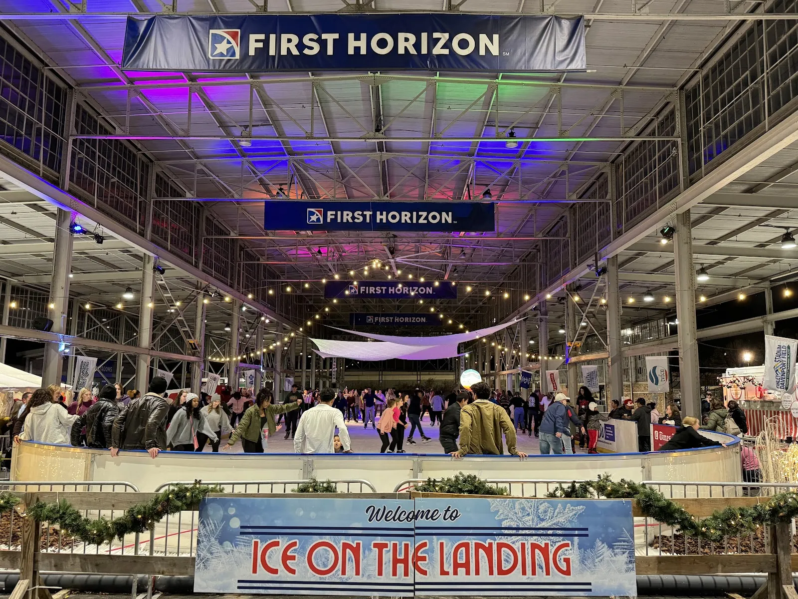 The covered First Horizon Pavilion with the Ice on the Landing welcome sign over the rink and skaters in the distance — Ice on the Landing