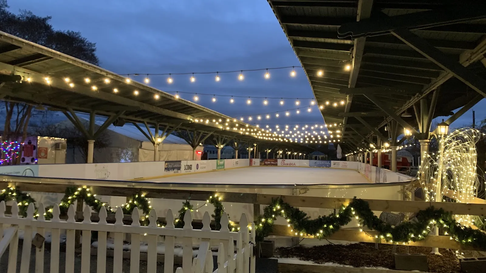 The fenced Ice on the Landing rink at dusk under string lights, with garland on the boards and no skaters on the ice — Ice on the Landing