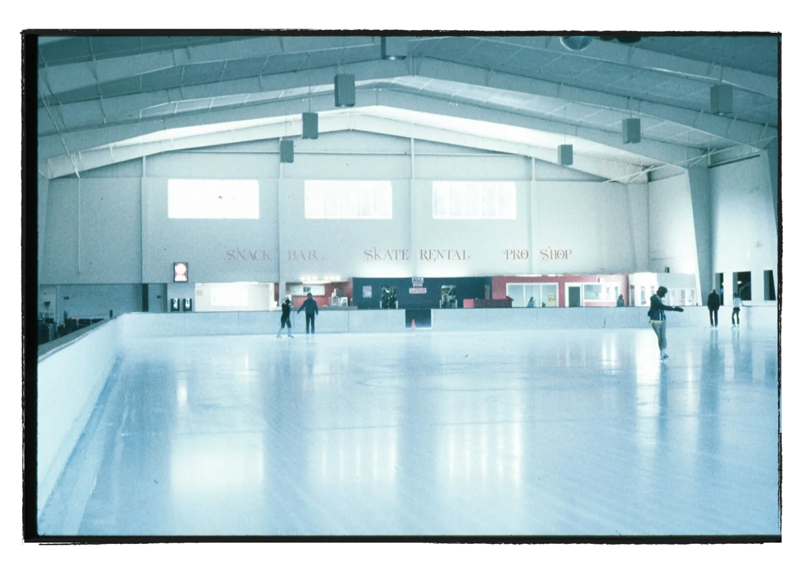 Most of the ice sheet from rink-side, with 'SNACK BAR SKATE RENTAL PRO SHOP' lettering on the back wall and a few skaters scattered on the ice. — Ice Palace Hawaii
