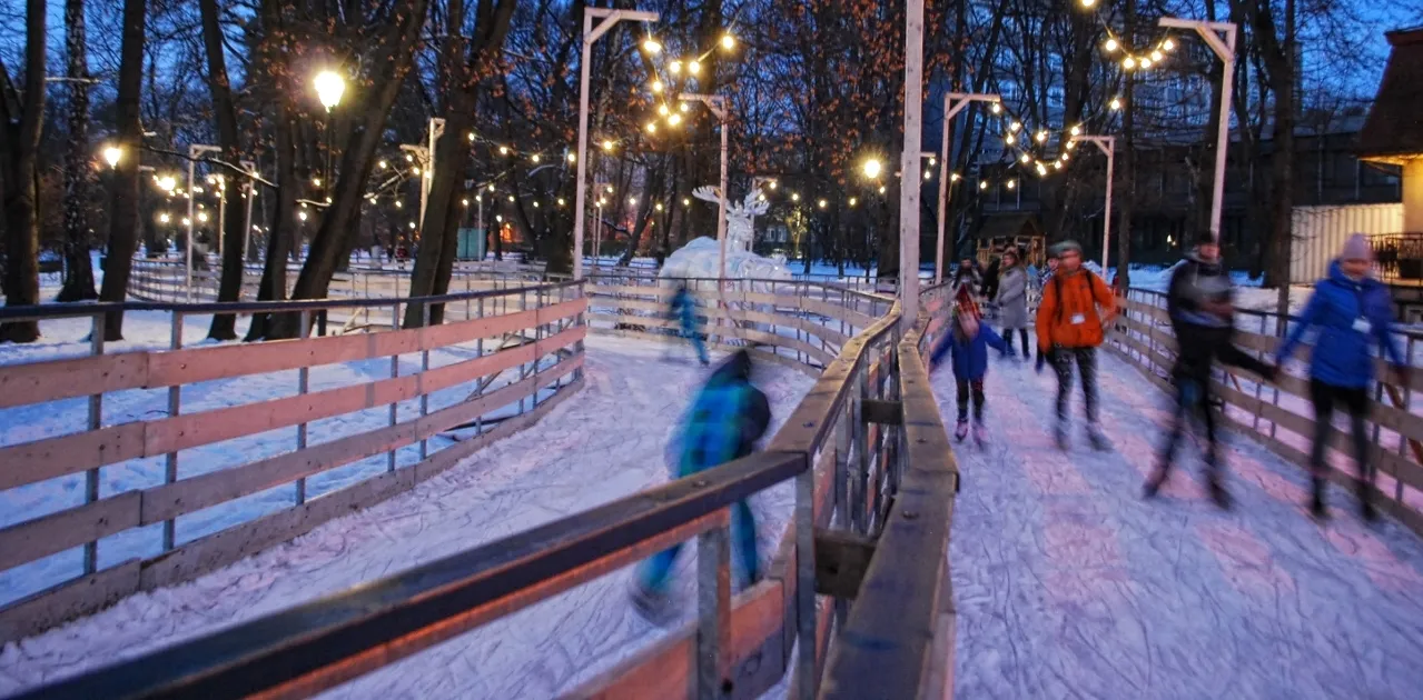 A lit ice path winding between trees in Park Jordana at dusk, with skaters along the boards. — Ice Park Kraków (Park Jordana)