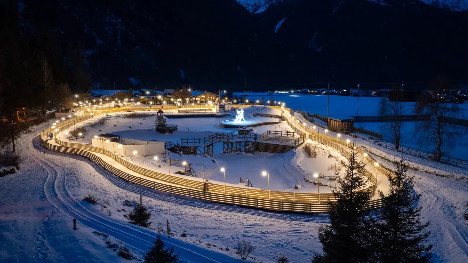Der beleuchtete Eisweg des Ice Parks Umhausen rund um den Naturbadesee im Winter, nachts von oben gesehen – Ice Park Umhausen
