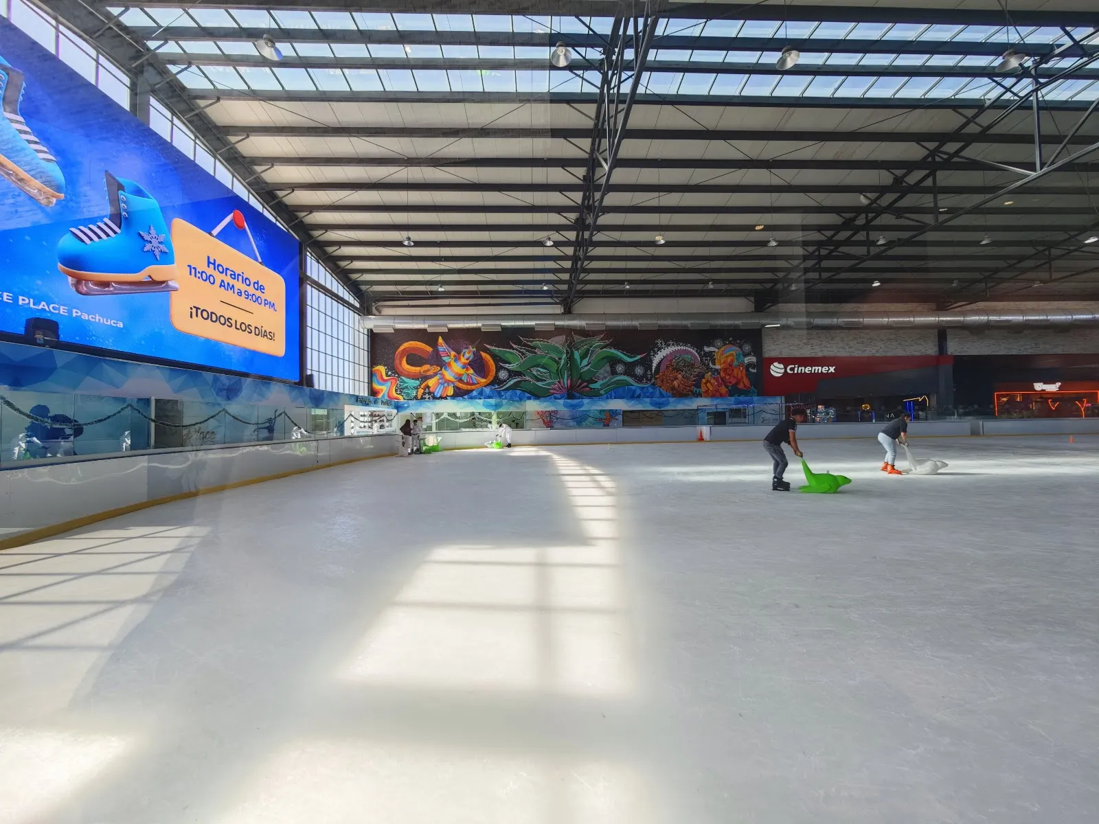 The wide ice of Ice Place at Explanada Pachuca under the skylit steel roof, with the rink banner and mural along the far wall and two children with skating aids on the ice — Ice Place Pachuca
