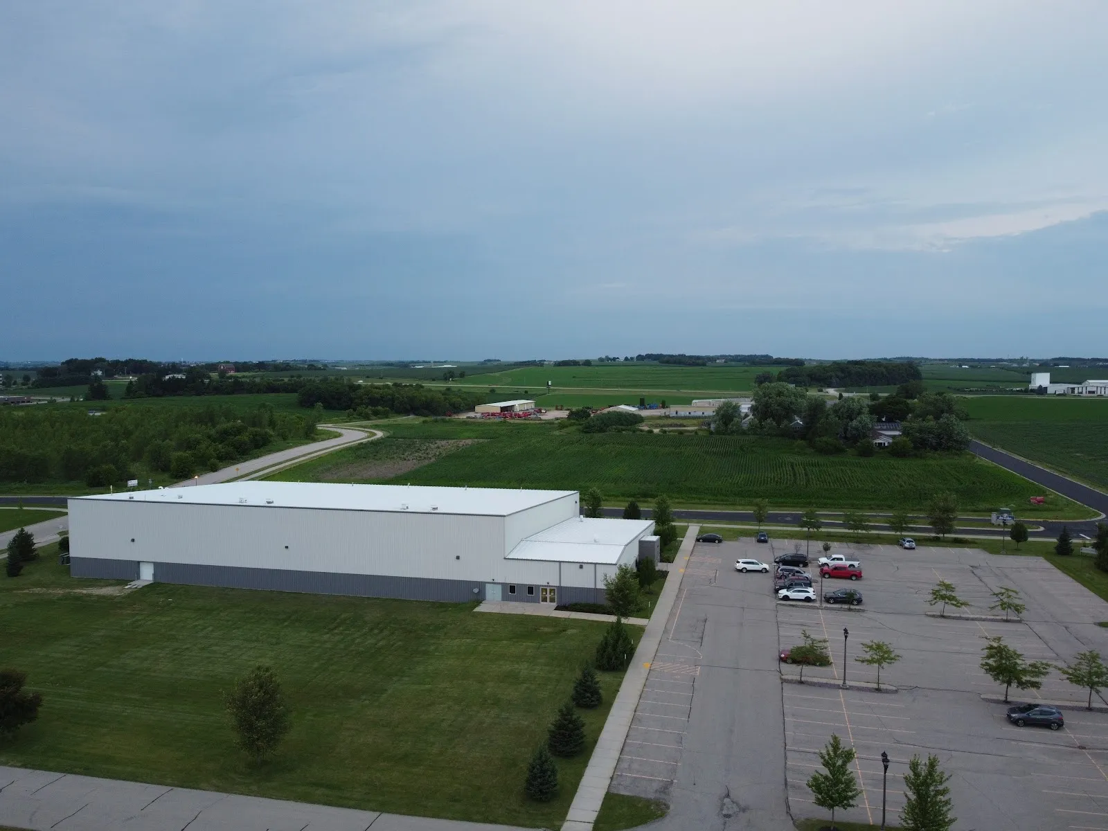 Aerial view of The Ice Pond at Waunakee: a long white metal-sided building with its parking lot beside it, set among green fields under a cloudy evening sky. — The Ice Pond at Waunakee