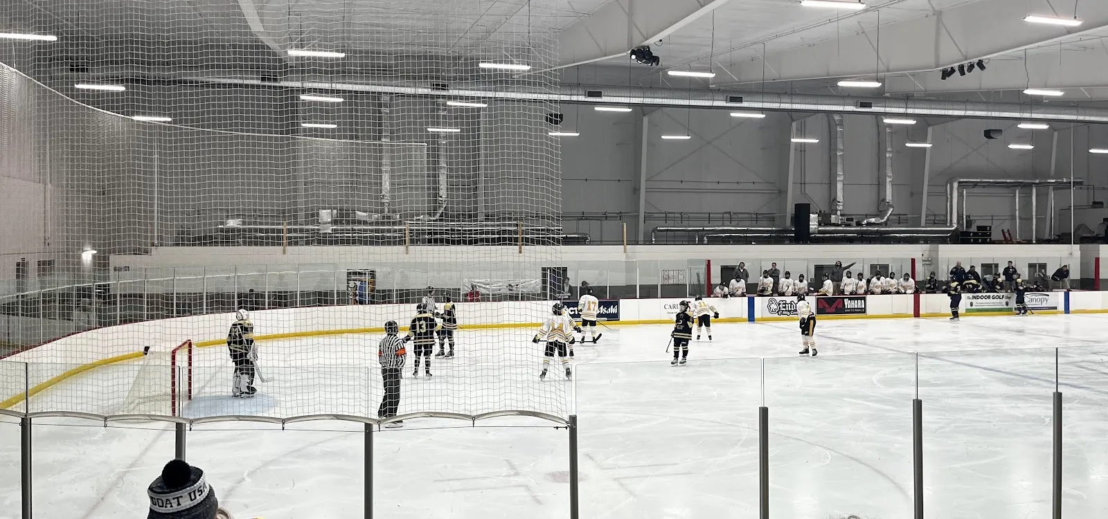 The ice from behind the glass under the bright white arched roof, with a youth game at the near end and sponsor boards along the far side. — The Ice Pond at Waunakee