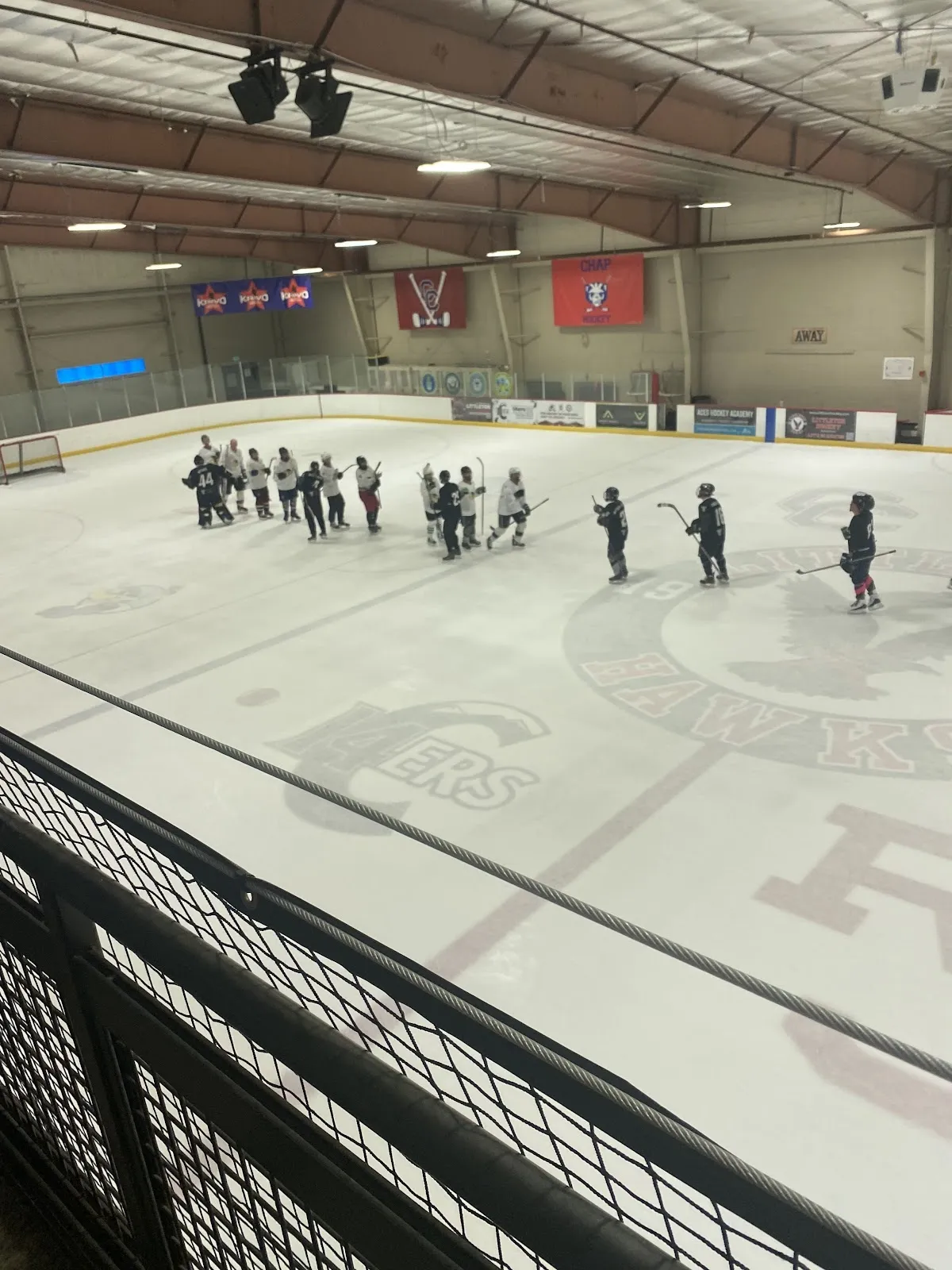 A wide view down onto the ice at The Ice Ranch from the rail, with a skills session at centre ice, the Hawks logo on the ice and the steel-trussed roof overhead. — The Ice Ranch